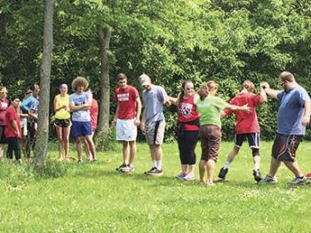 A group of teens and adults stands in a grassy field holding hands or linking arms, working together in a team-building activity, with trees and greenery in the background.