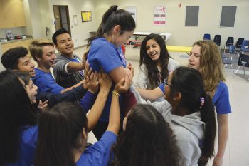 a group of students with hands raised hold up another student with her arms crossed