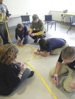 A small group of teens and adults kneels on a classroom floor, working together to measure and mark lines with yellow tape, collaborating on a hands-on activity with chairs visible in the background.