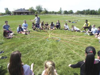A group of kids sits in a large circle on a grassy field, each holding a piece of string connected to the center, creating a web-like pattern during a team-building activity, with a building and trees in the background.