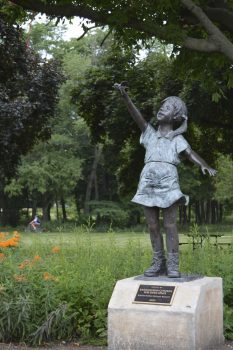 A statue of a young girl standing on a pedestal reaches upward, surrounded by greenery and flowers in a park setting.