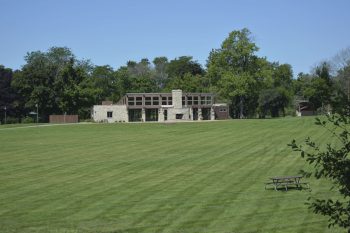 A wide grassy field stretches toward a building in the distance, with trees lining the area and a picnic table visible in the foreground.