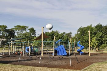 A playground with climbing structures, swings, and blue slides sits in a park, surrounded by trees under an open sky.