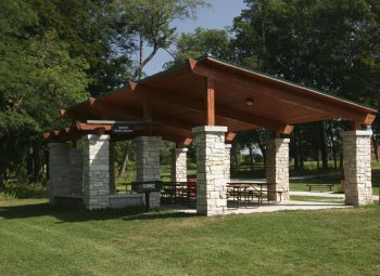 A covered picnic pavilion with stone columns and a wooden roof sits in a grassy park area surrounded by trees.