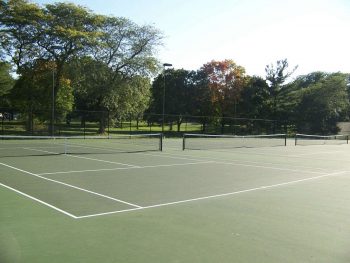 An outdoor tennis court with painted lines sits empty, surrounded by trees and greenery under a clear sky.