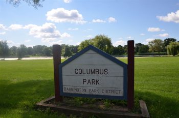 Columbus Park sign on the edge of an open field adjacent to a lake. Reads Columbus Park, Barrington Park District