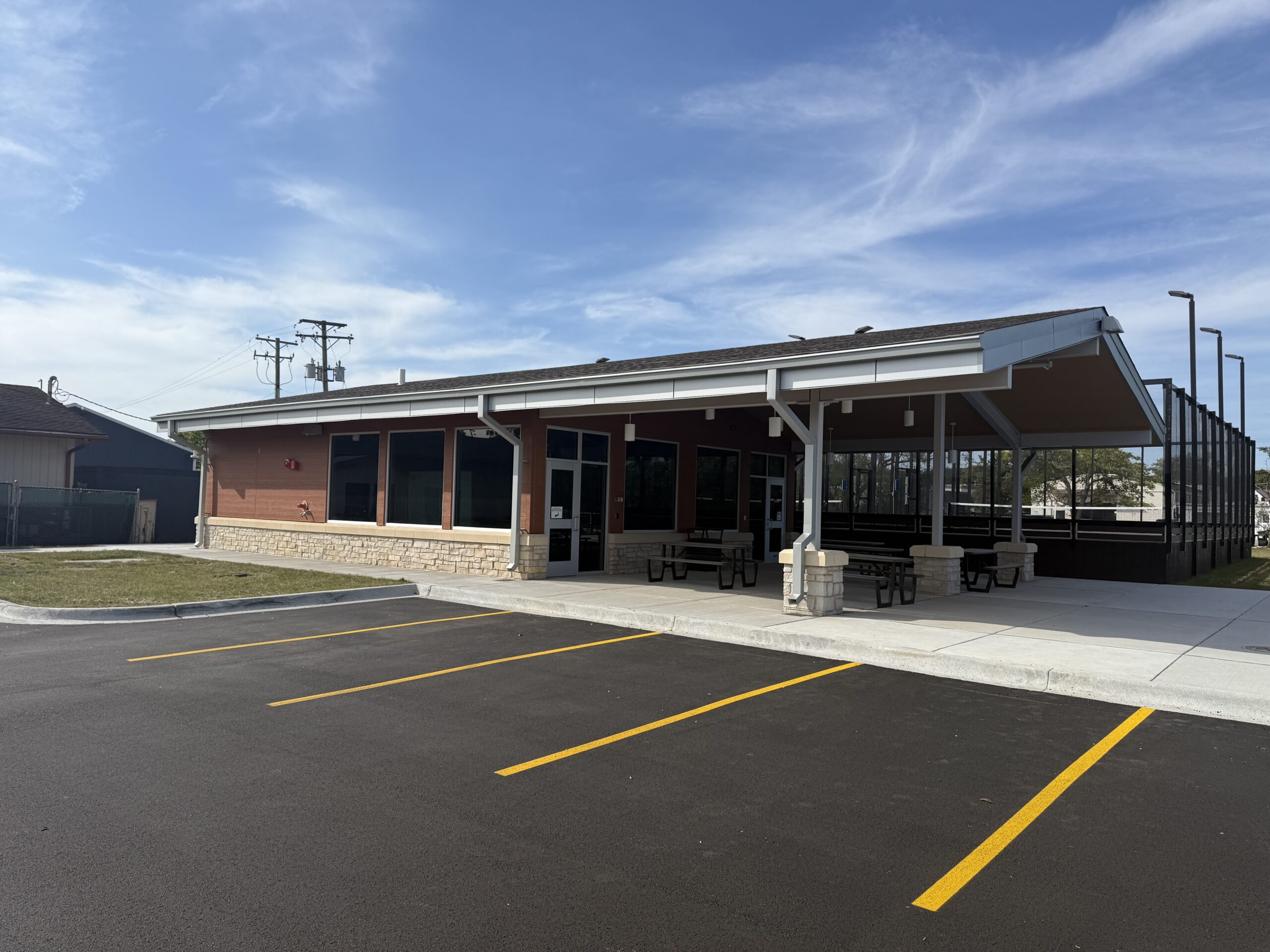 Recreation building with covered patio seating and adjacent outdoor courts next to a parking lot.