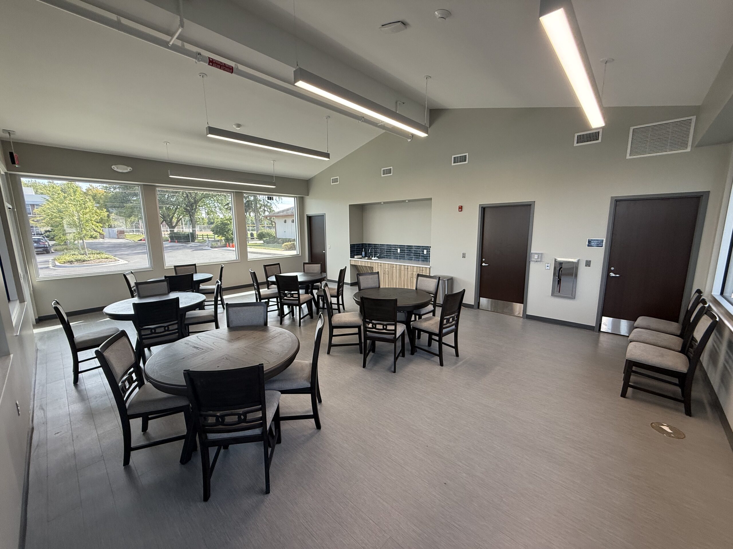 Indoor community room with round tables and chairs, large windows, and modern lighting.