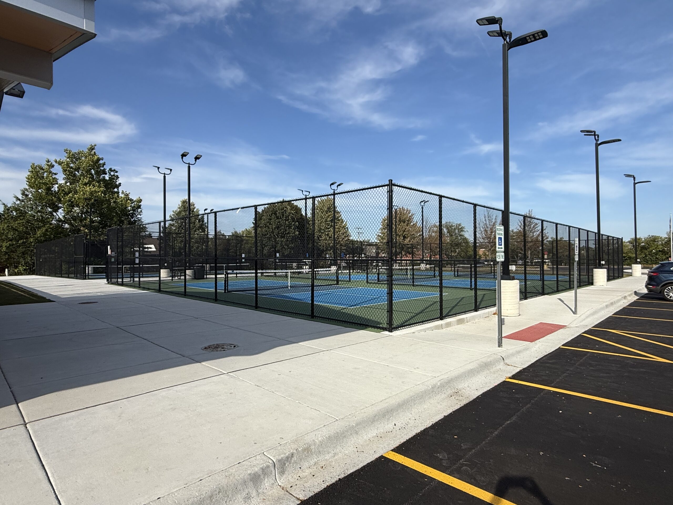 Outdoor pickleball courts enclosed by fencing next to a parking lot and sidewalk.