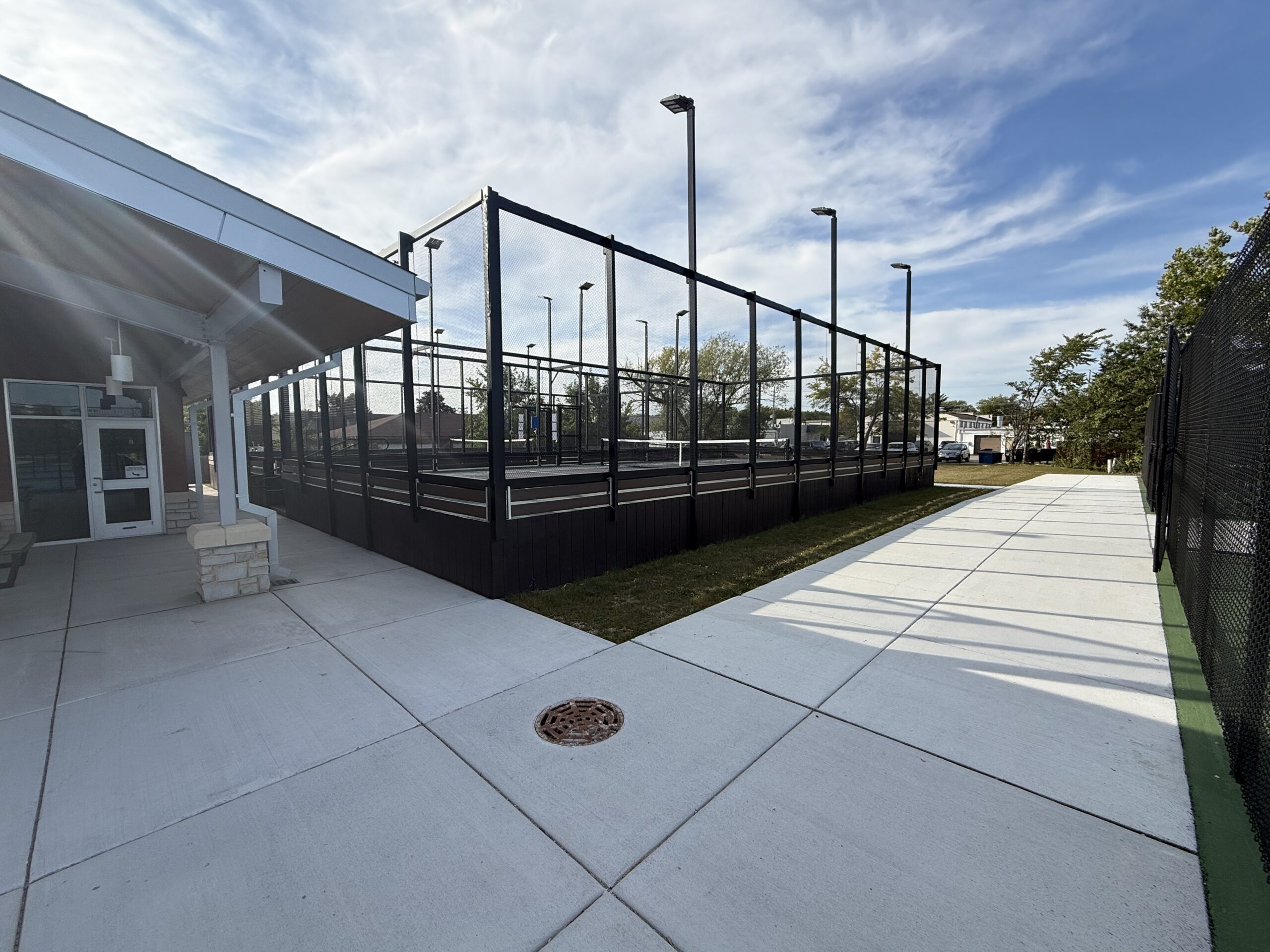 Outdoor tennis courts with heated flooring enclosed by black fencing next to a recreation building and sidewalk.