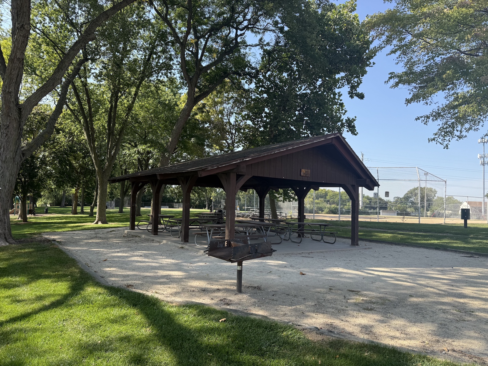 Picnic shelter with wooden pavilion, picnic tables, and grill in a shaded park near a baseball field.