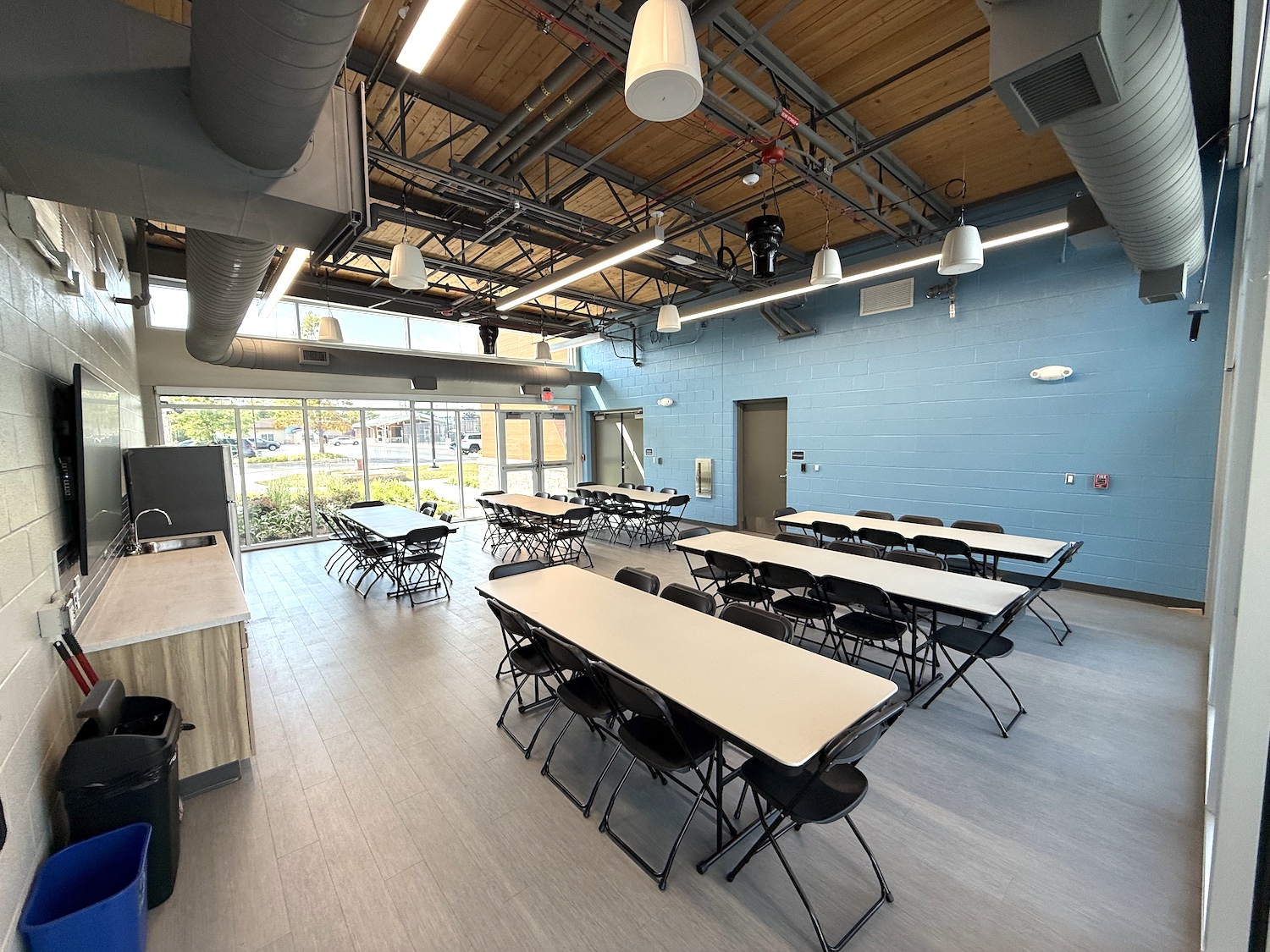 A bright, modern meeting room with tables and folding chairs arranged in rows, featuring large windows, blue walls, and exposed ceiling ducts.