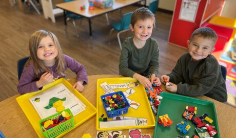 Three young children sitting at a classroom table, smiling while playing with colorful building toys and learning materials on trays.