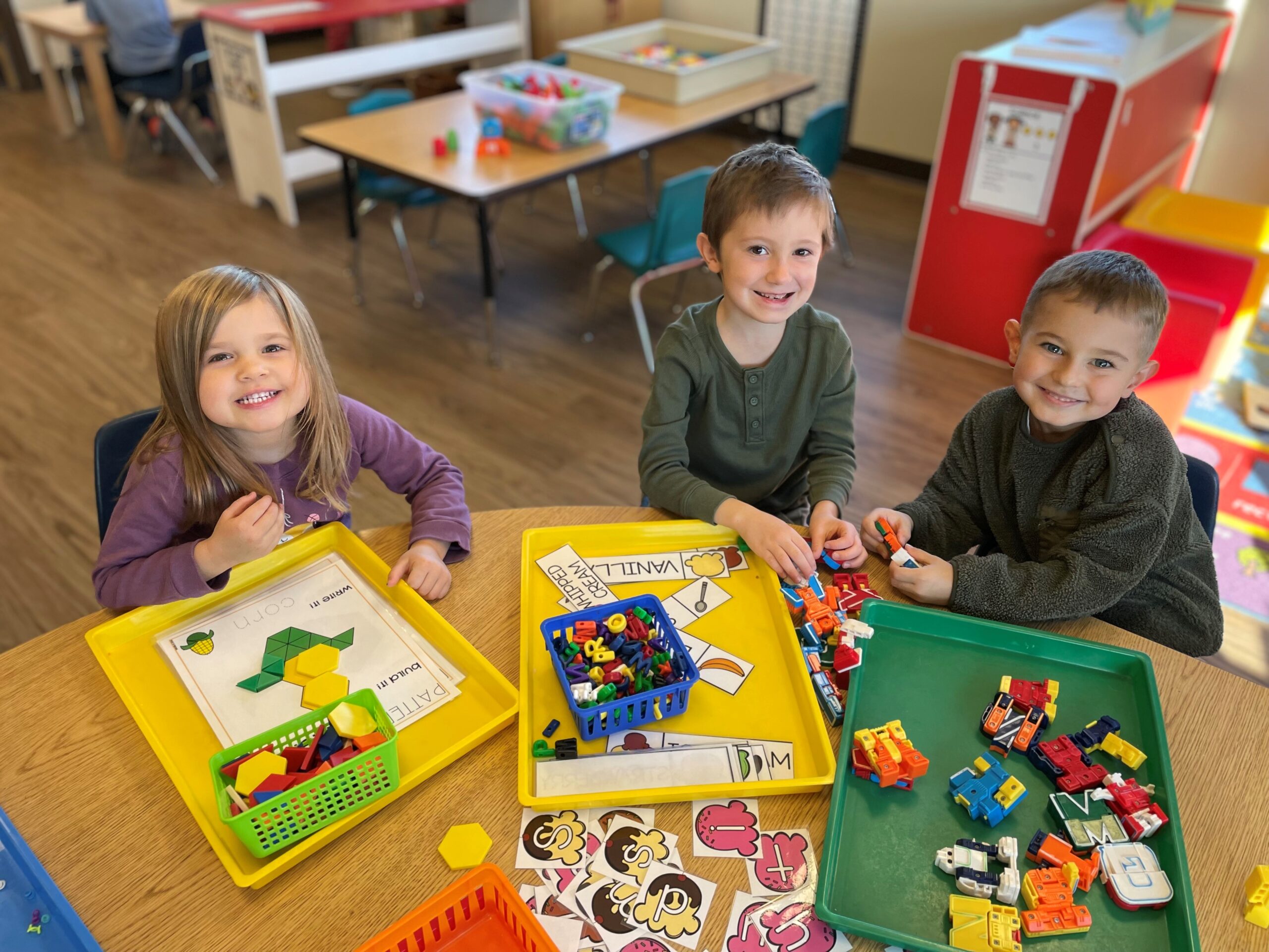A group of three young children smile for the camera while seated at a table in a preschool classroom with various activities on the table.