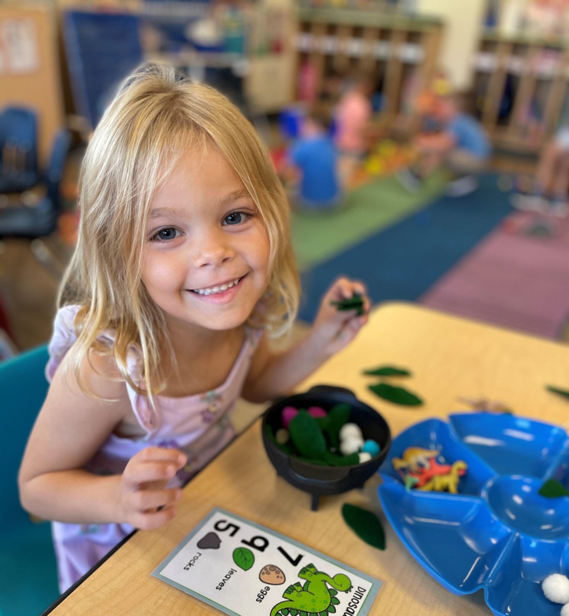 A young girl in a preschool classroom poses for the camera while sitting at the table playing with dinosaur toys.