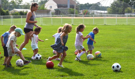 A group of young children dribble soccer balls across a grassy field while following an instructor during an outdoor youth sports class.