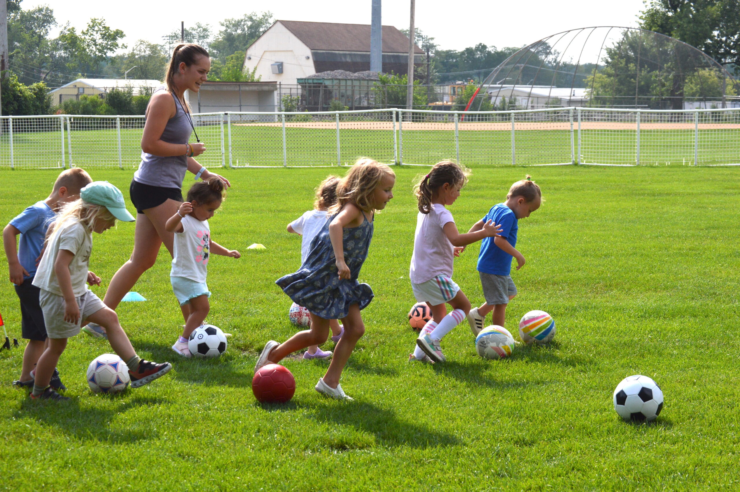 A group of young children run across a grassy field kicking soccer balls, while an adult instructor walks alongside them during an outdoor activity.
