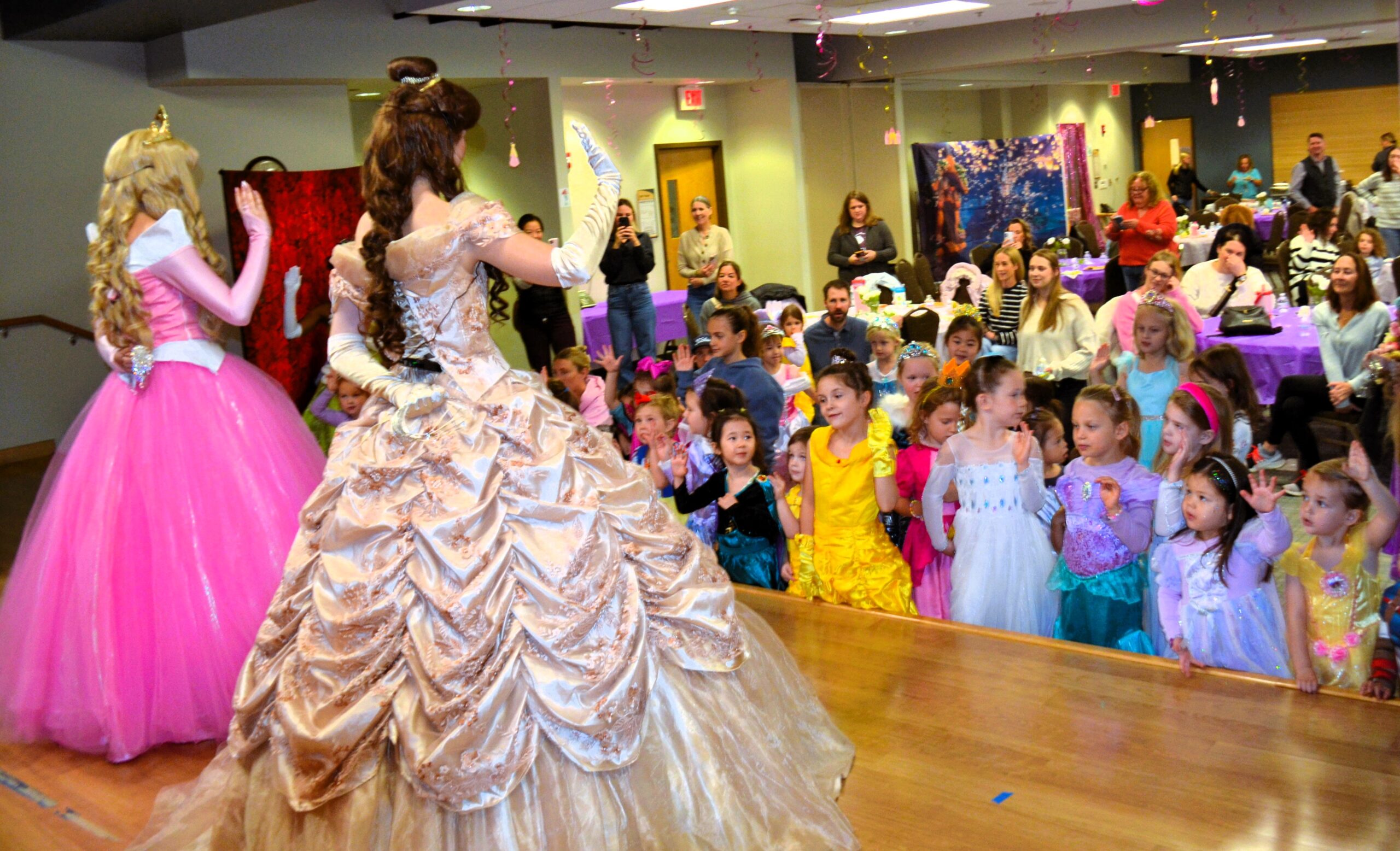 Two costumed princess performers stand on a stage leading an activity, while a group of young children watch and follow along at a party.