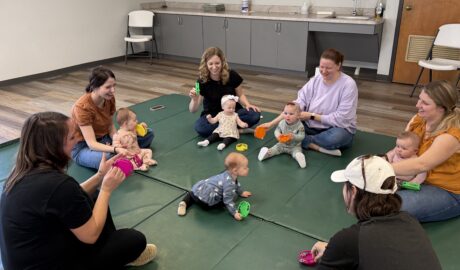 Caregivers and babies sitting in a circle on a padded floor during a parent-child class, with adults smiling and engaging infants using colorful toys.