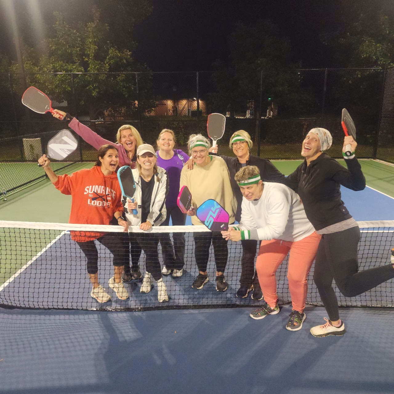 A group of adults stand together on an outdoor pickleball court at night, smiling and holding paddles as they pose playfully near the net.