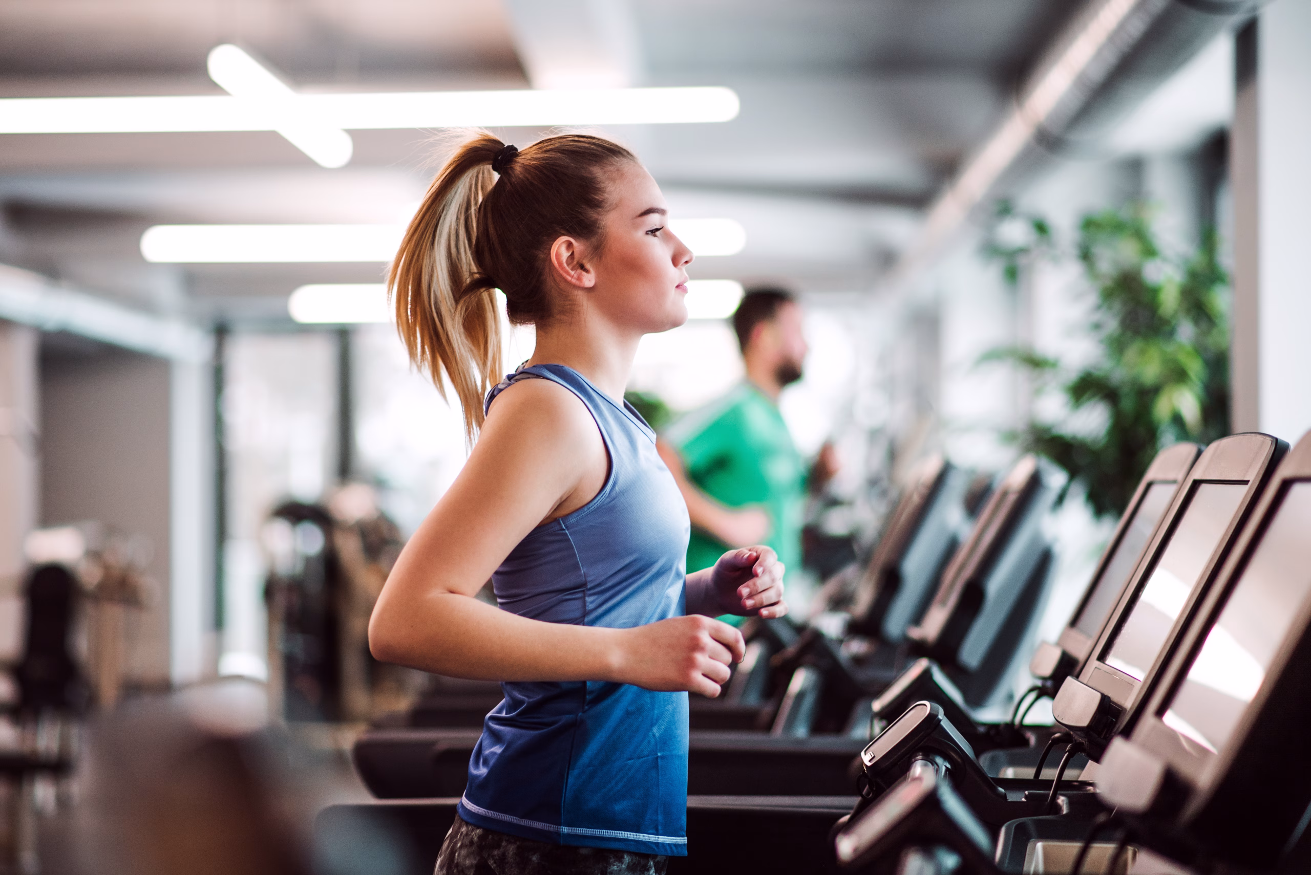 A woman jogs on a treadmill in a gym, with other exercise equipment and people working out in the background.