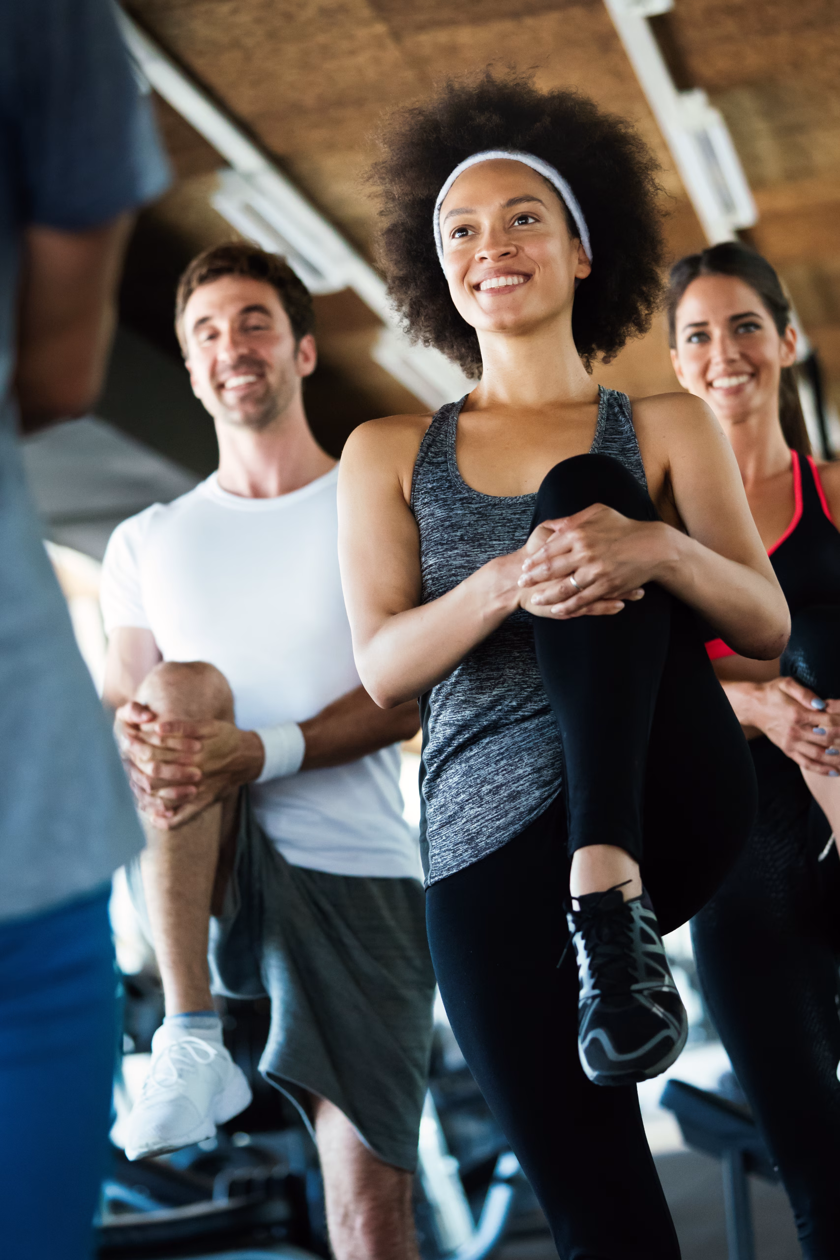 A small group of adults stretch during a fitness class, balancing on one leg while holding their knee, smiling in a gym setting.