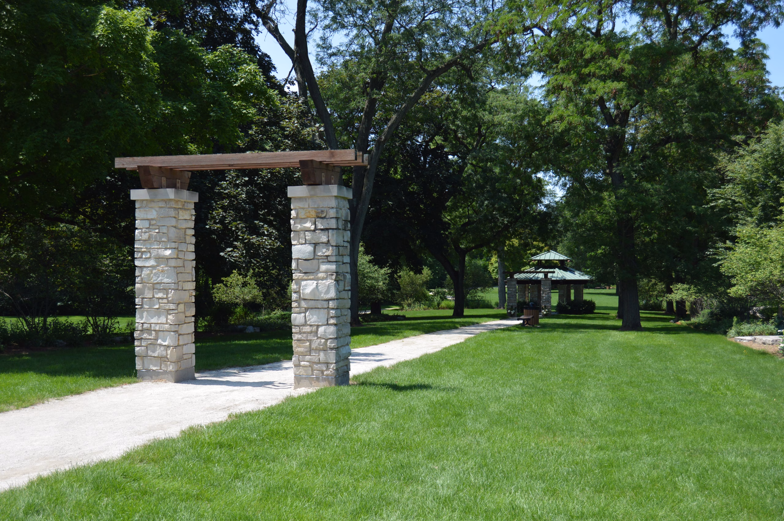 A scenic park pathway lined with green grass and tall trees passes under a stone archway, leading toward a small gazebo in the distance.