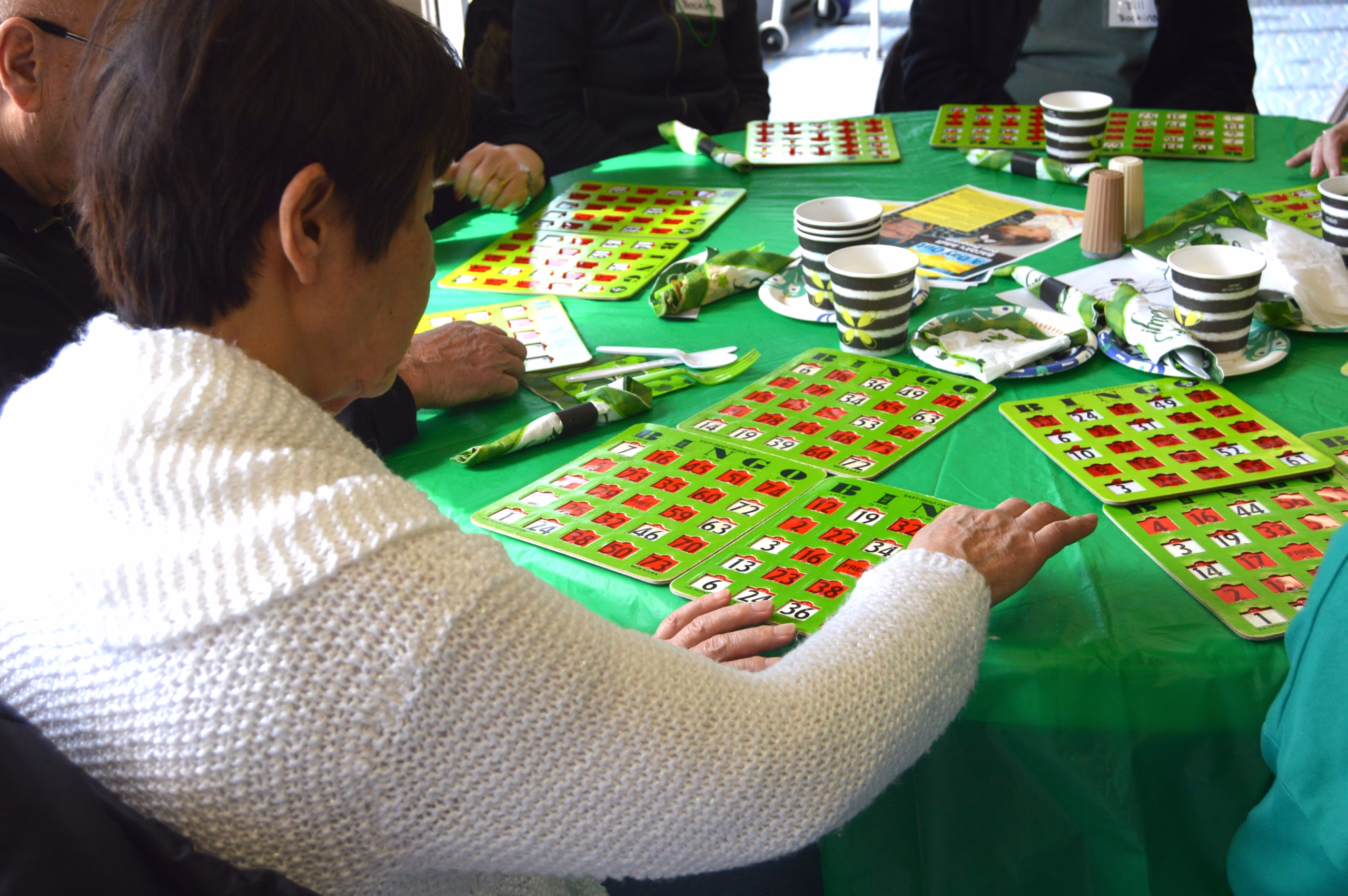 A group of older adults sit around a table playing bingo. Several cards, cups, napkins, and game markers are spread across the table.