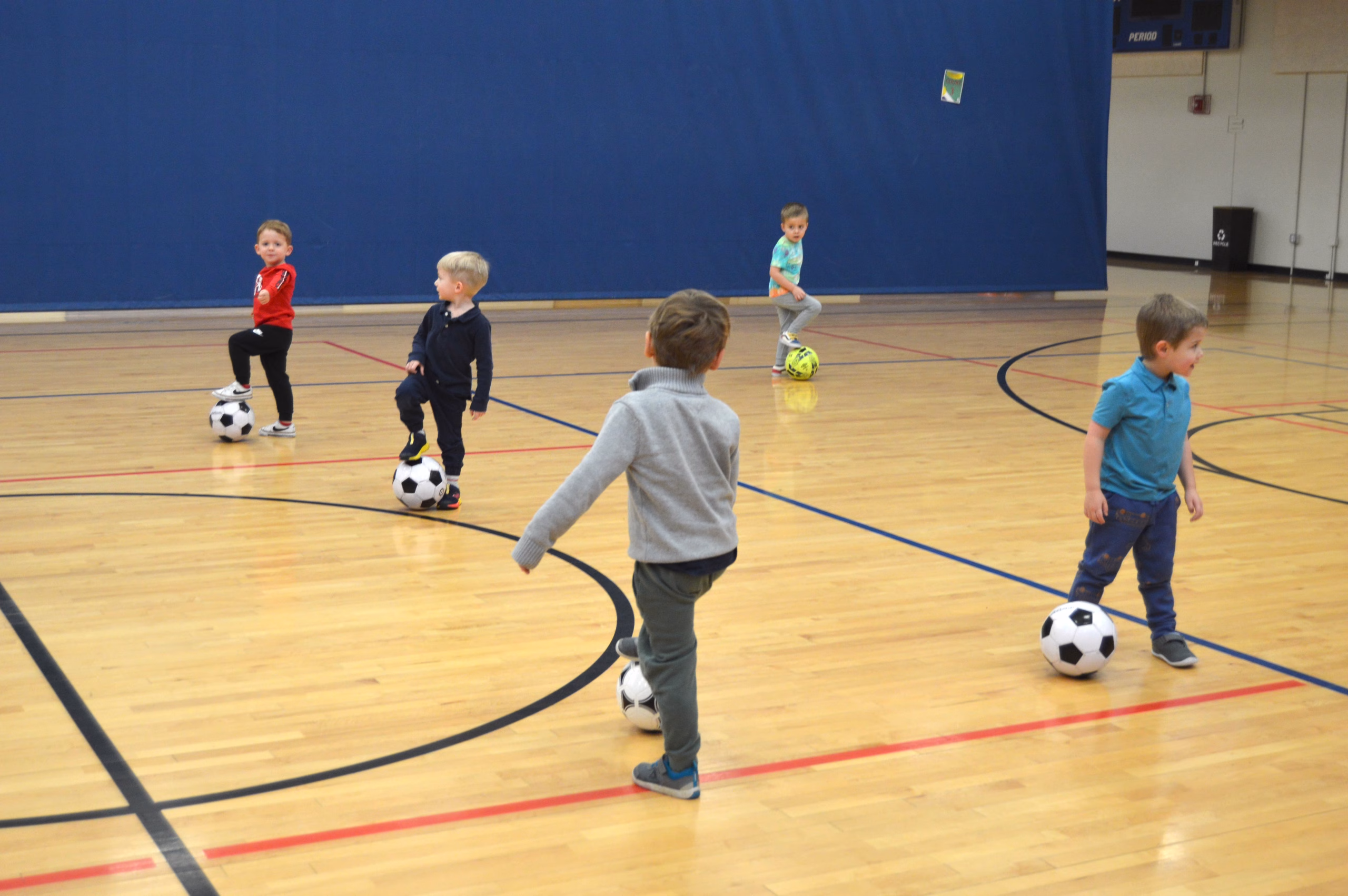Several young children play with soccer balls on an indoor gym floor, moving and practicing basic skills during a group activity.