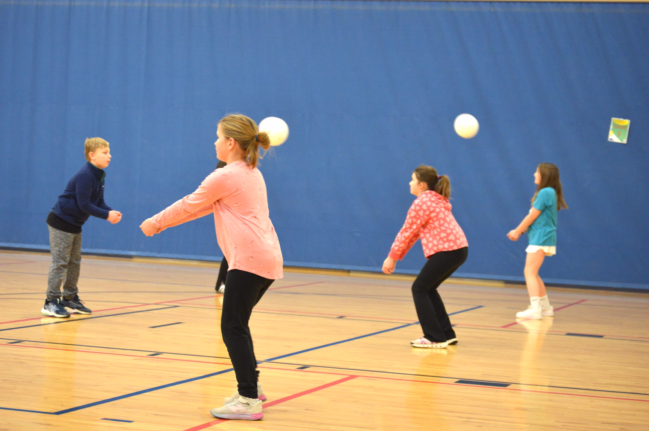 A group of children stand in a gym preparing to pass or hit a ball, practicing a team activity with multiple balls in motion against a blue wall background.