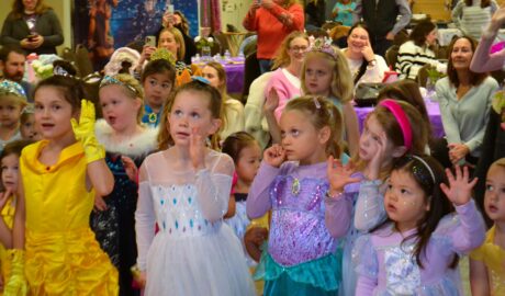 A group of young children dressed in princess costumes raise their hands and follow along during an indoor party activity, while adults watch and take photos in the background.