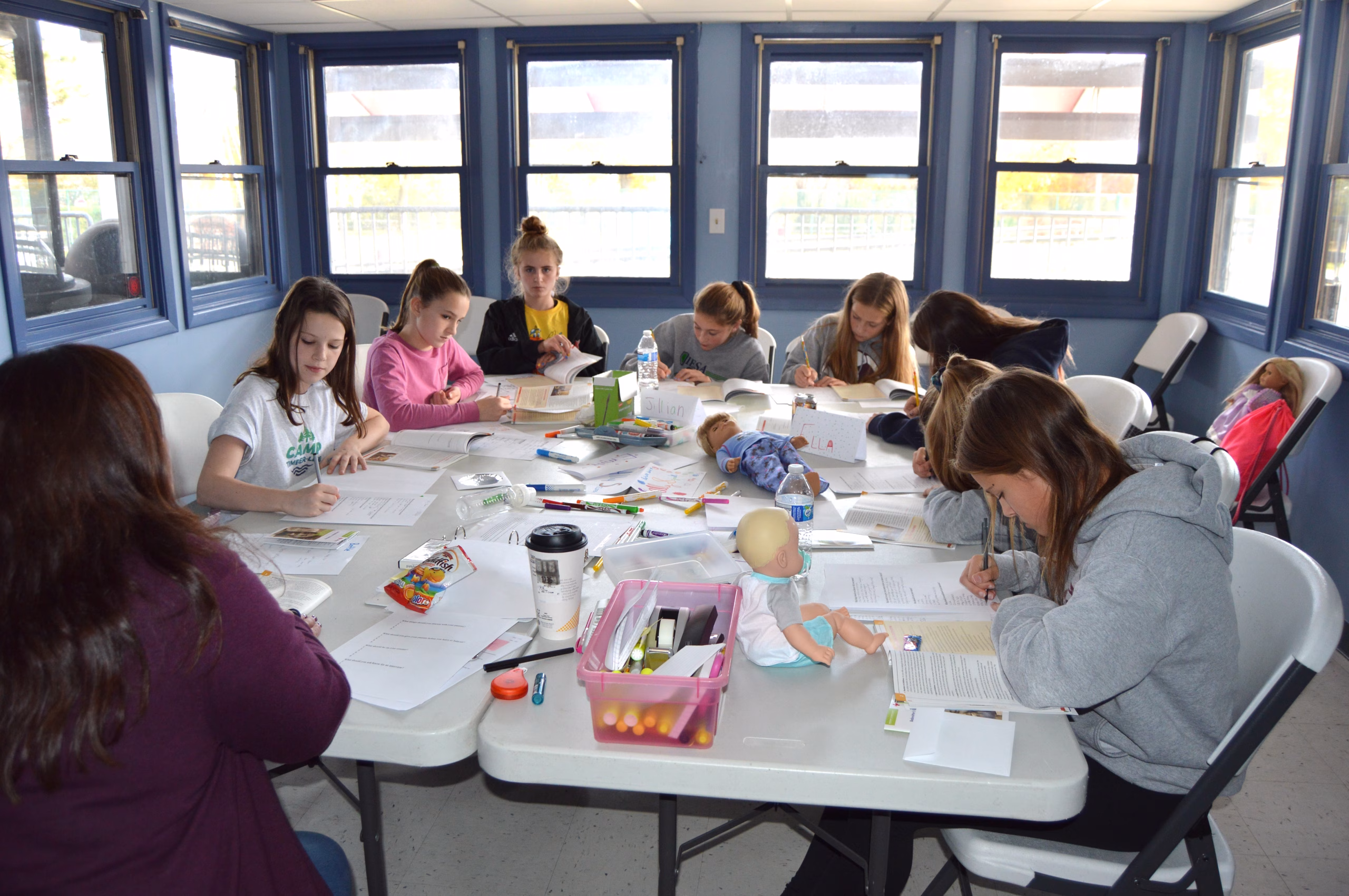 A group of teens sit around a table focused on writing and completing worksheets, with school supplies and materials spread out in front of them.