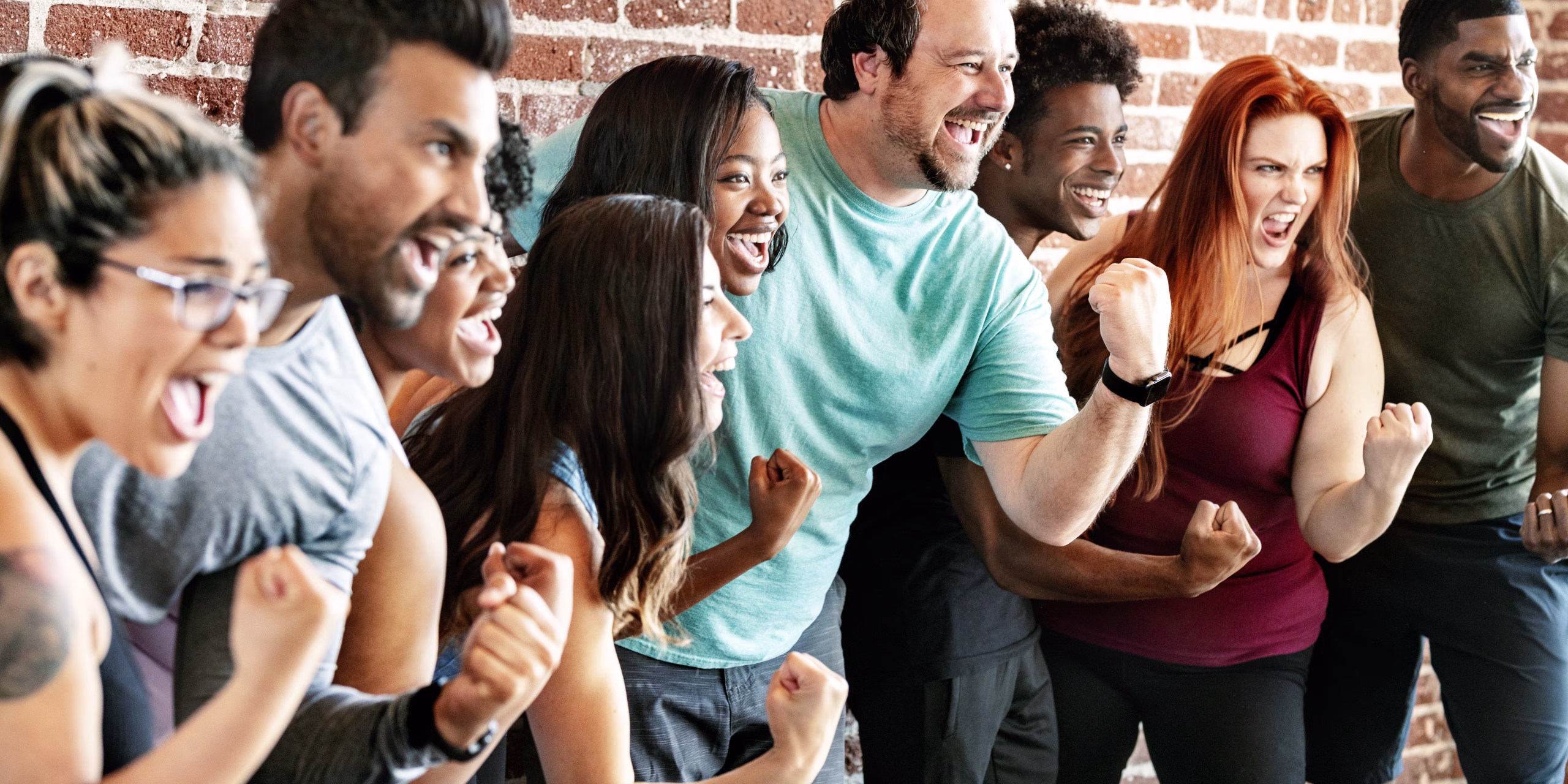 A diverse group of adults stand together indoors, cheering and smiling with raised fists, appearing excited and celebratory.