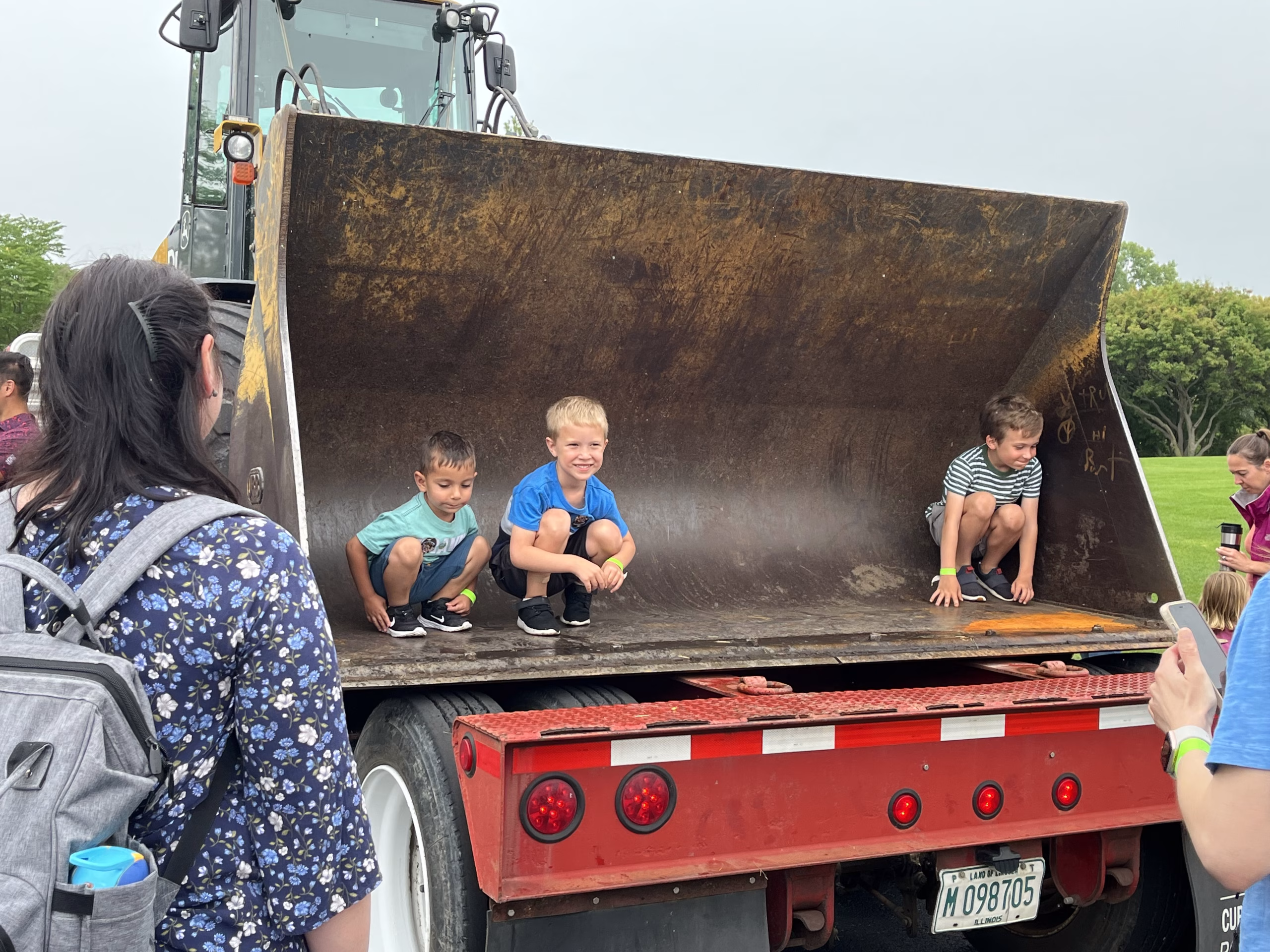 Three young children crouch inside the large bucket of a construction loader attached to a truck, while adults stand nearby watching and taking photos outdoors.