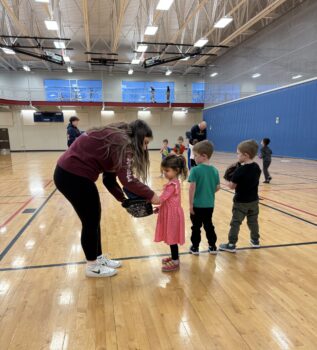 An instructor hands an item to a young child standing in line during a group activity in an indoor gymnasium, with other children waiting nearby.