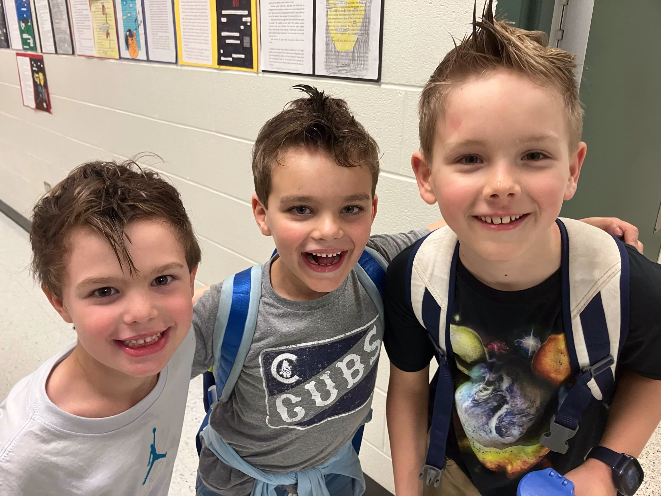 A group of three young boys in school wearing backpacks smile for the camera.