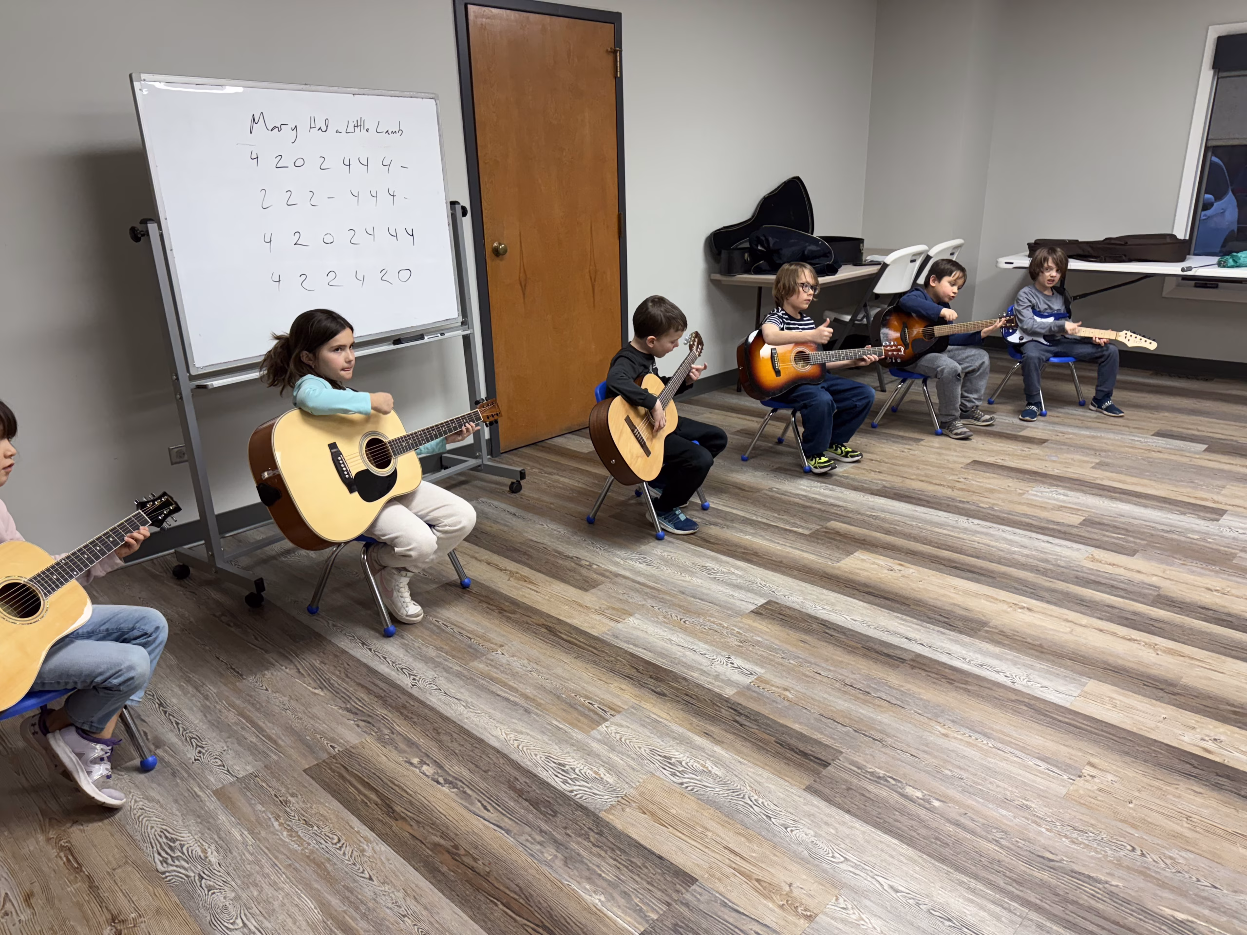 Young children sitting in chairs sit in a row holding their guitars. A white board in the background shows them the notes to play Mary had a Little Lamb.