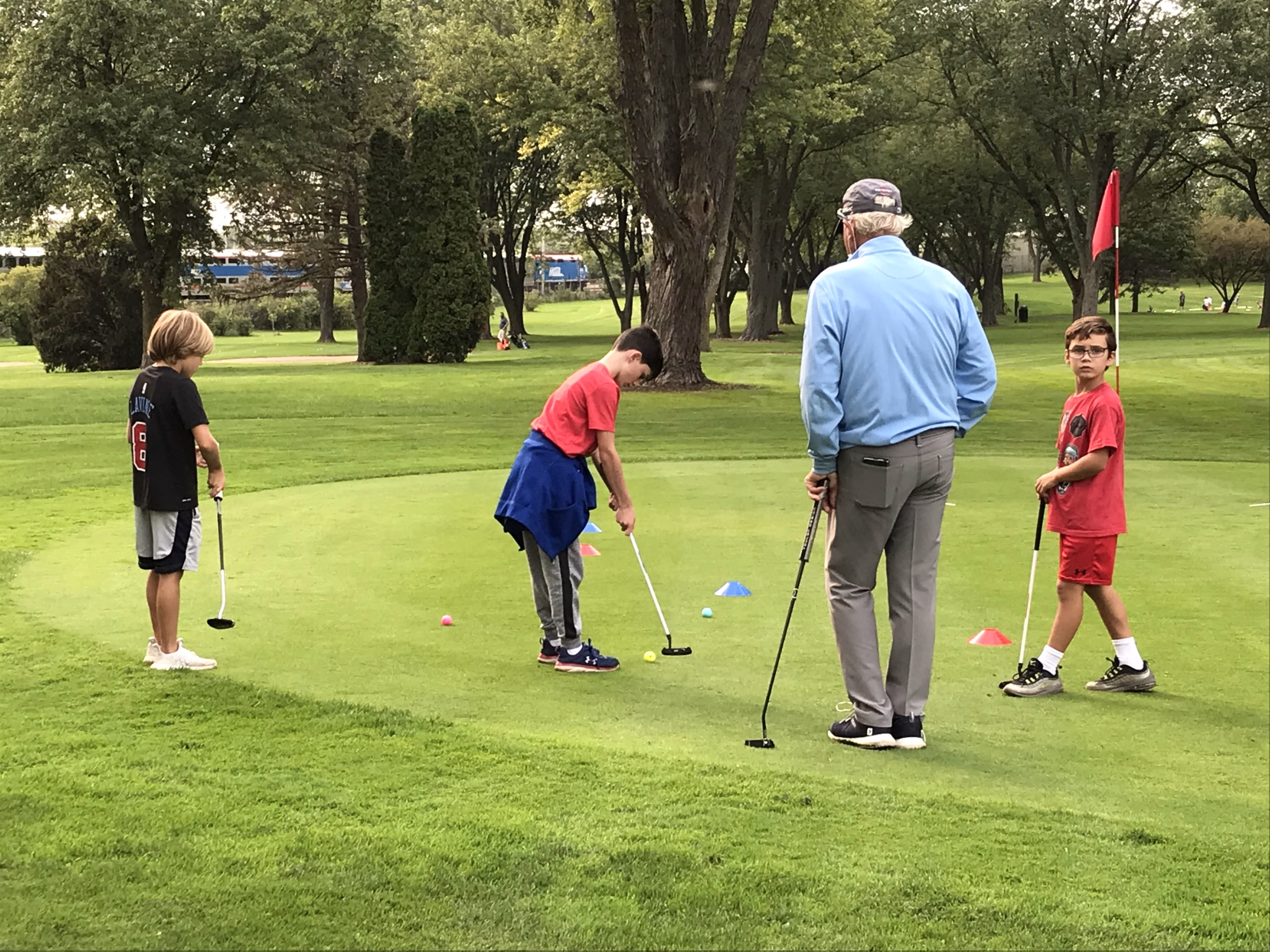 Three children practice putting on a golf green while an older instructor stands nearby, guiding them during a lesson on a tree-lined course.