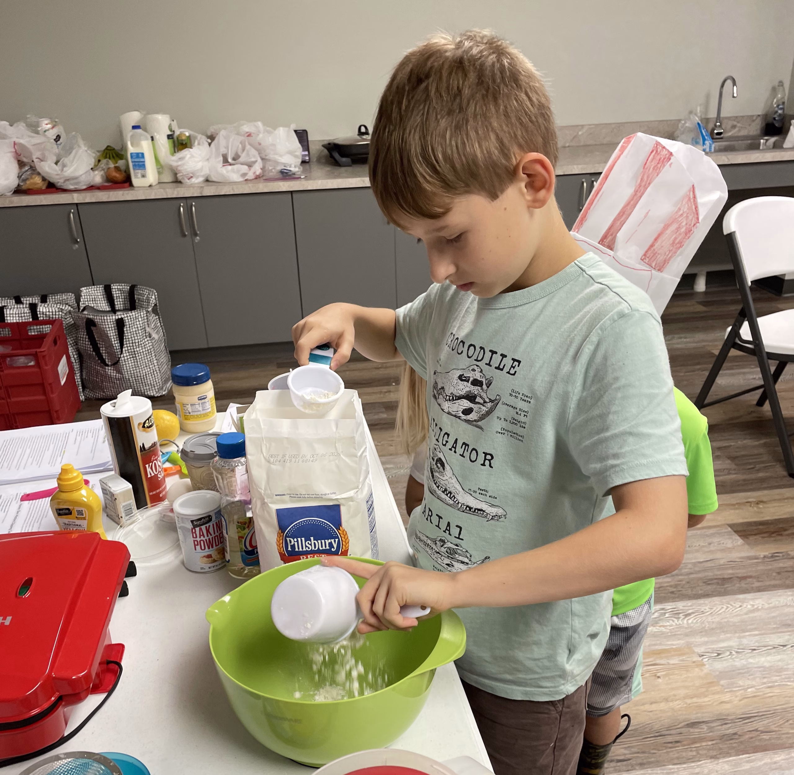 A young boy scoops flower with a measuring cup from a bag, and pours it into a green bowl on the counter with other baking ingredients near by.