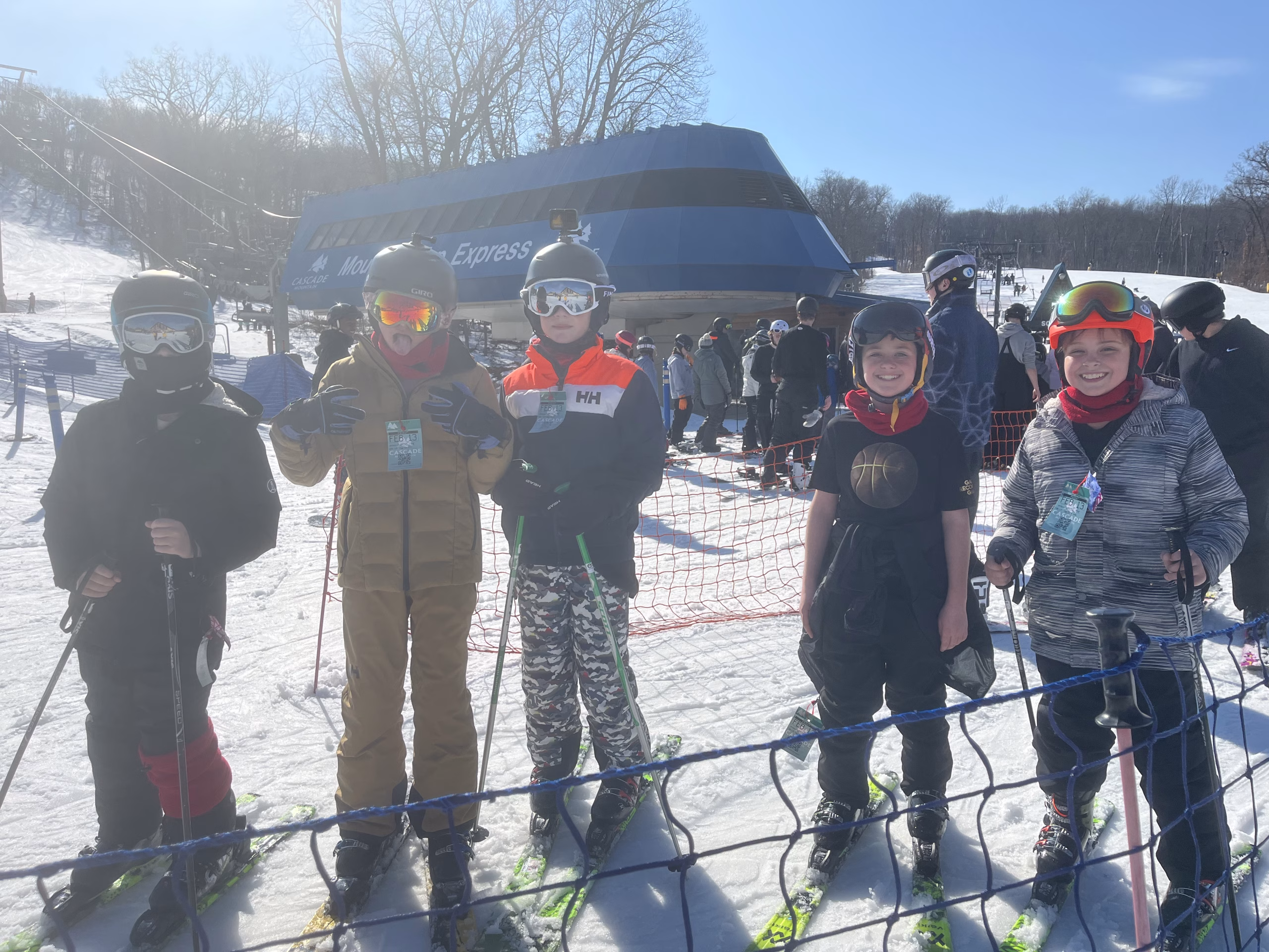 Five children wearing ski gear stand in a line holding ski poles and smiling in front of a busy ski lift area with other skiers in the background.