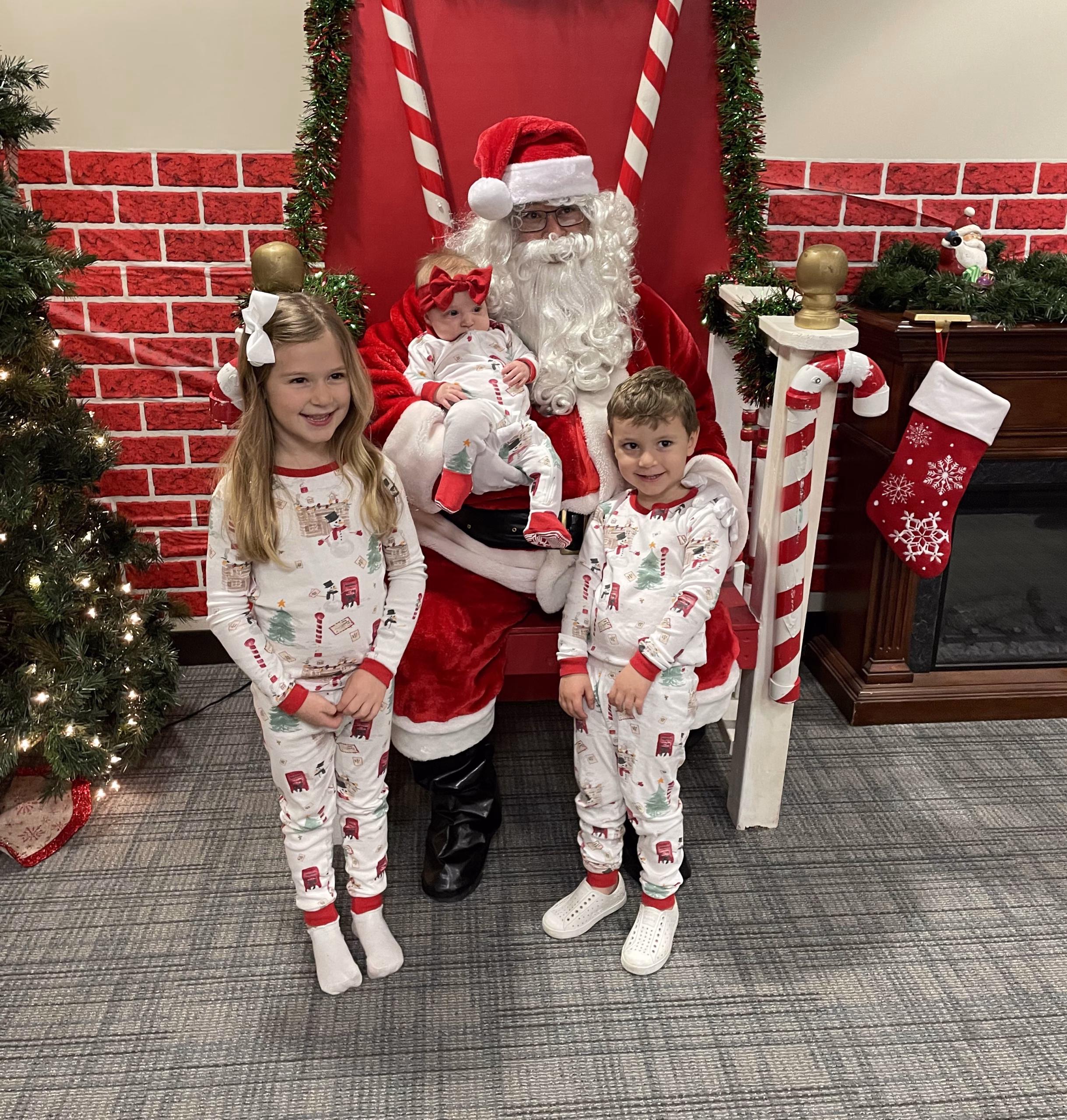 Santa, sitting in a chair holding a baby, while two siblings stand in front smiling for the camera.