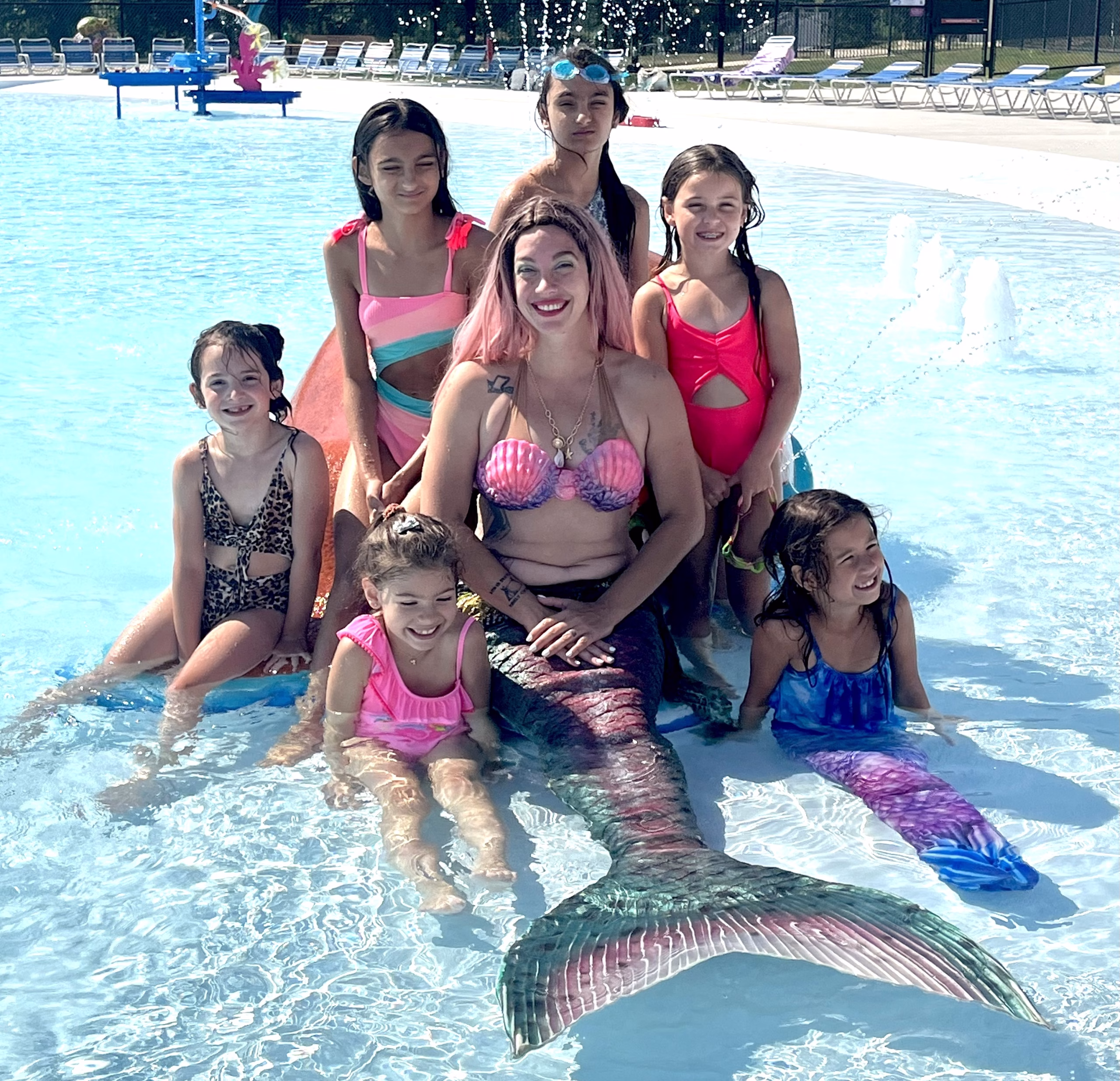 A person dressed as a mermaid sits in a shallow pool surrounded by smiling children in swimsuits, posing together in the water at an outdoor aquatic center.