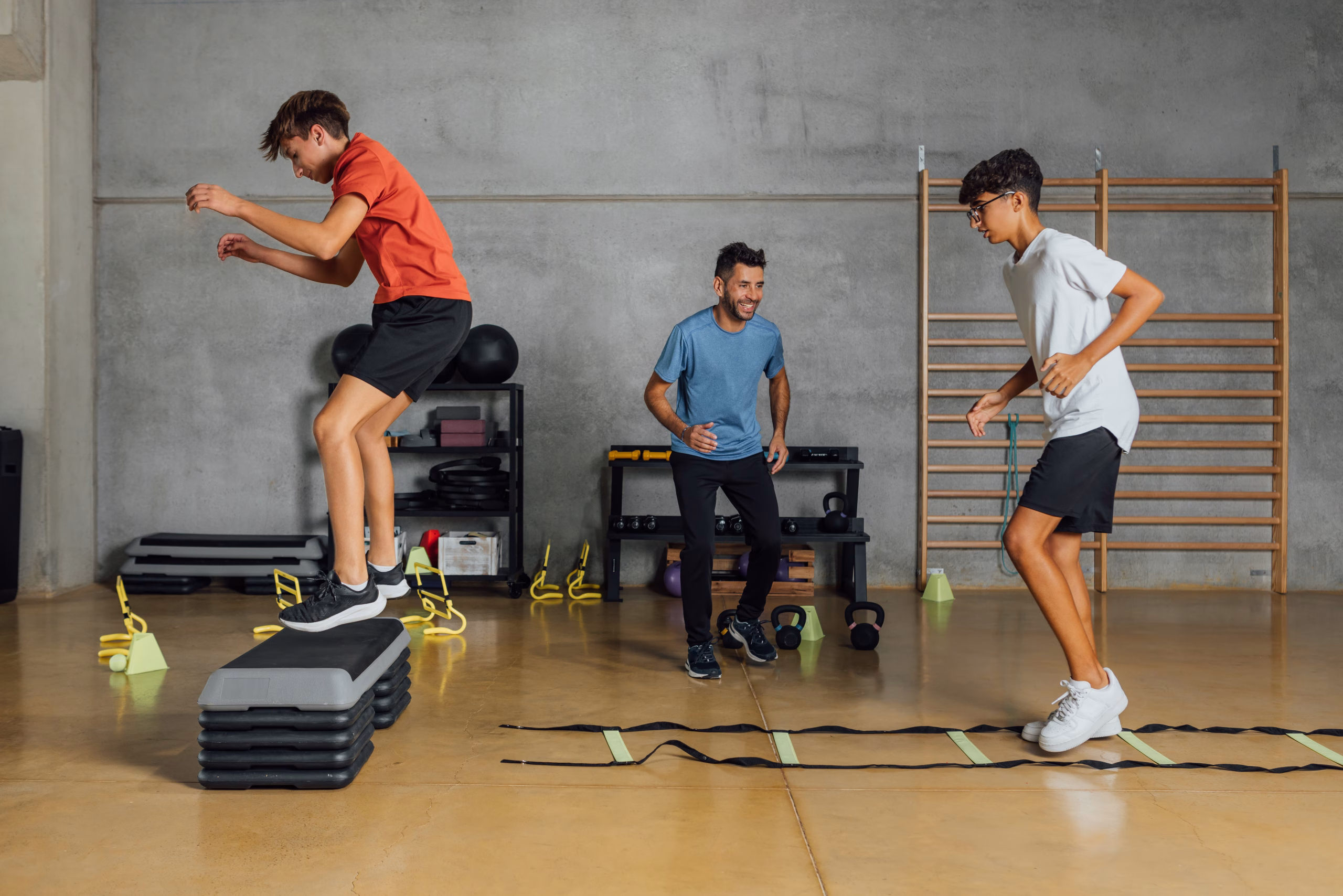 Two boys participate in an indoor training session, jumping and stepping through agility exercises while an instructor guides them in a gym setting.