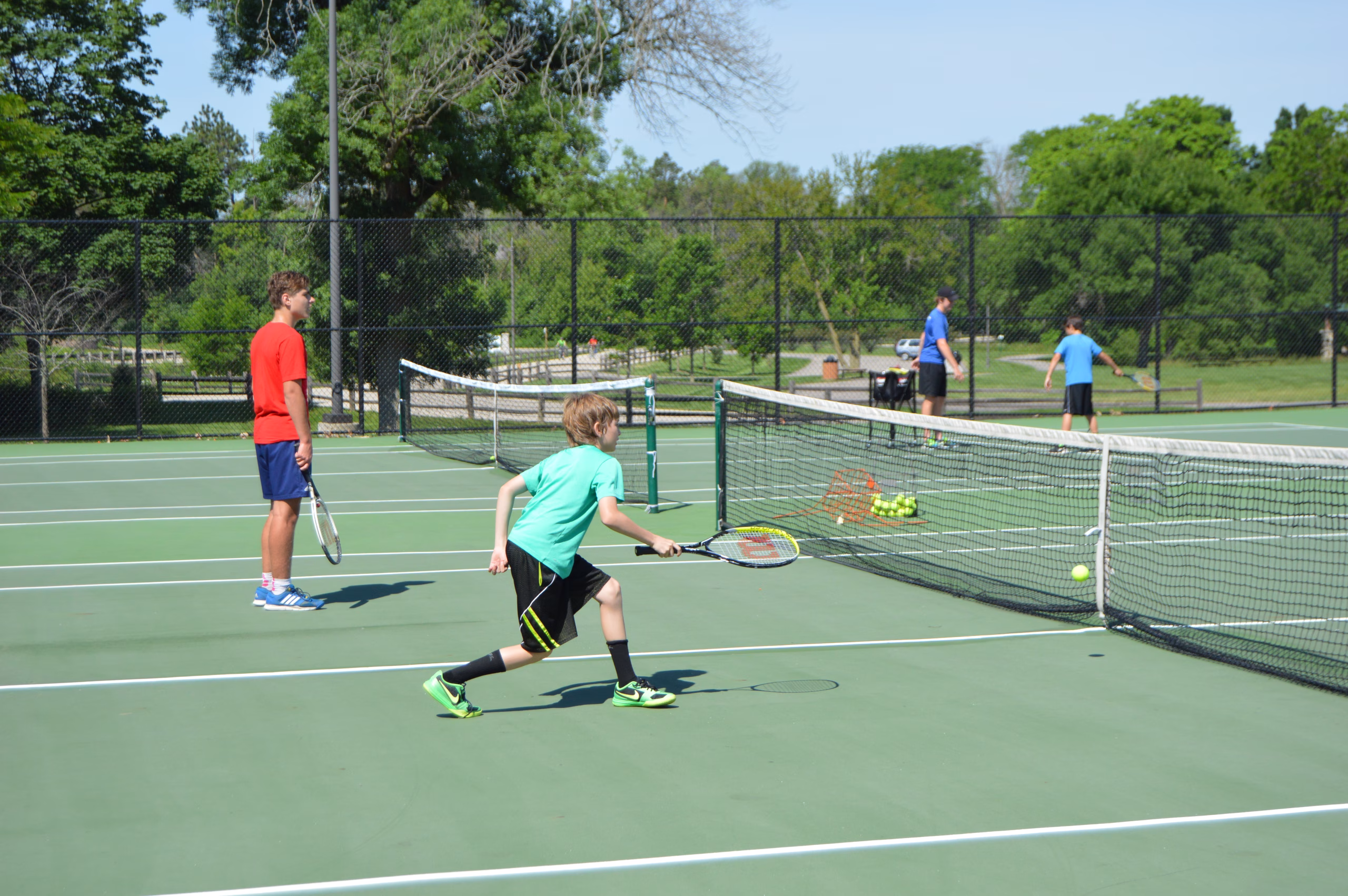 A child runs toward a tennis ball on an outdoor court while holding a racket, as an instructor and other players practice in the background.