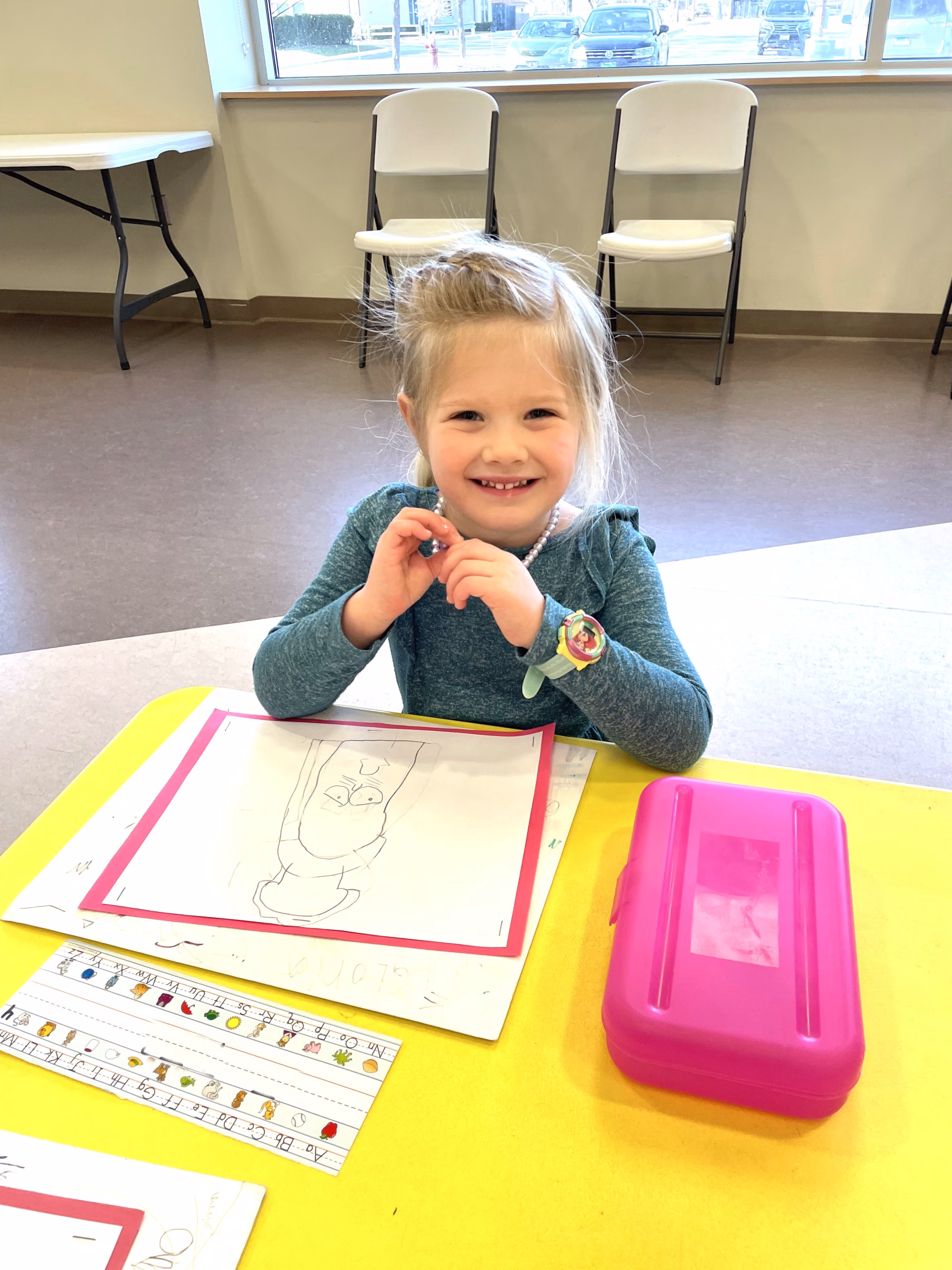 A little girl smiles for the camera while sitting at a table drawing.