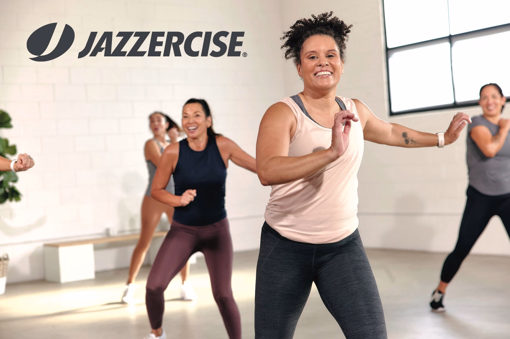 A group of women participate in a Jazzercise class, following an instructor as they move energetically in a bright fitness studio.