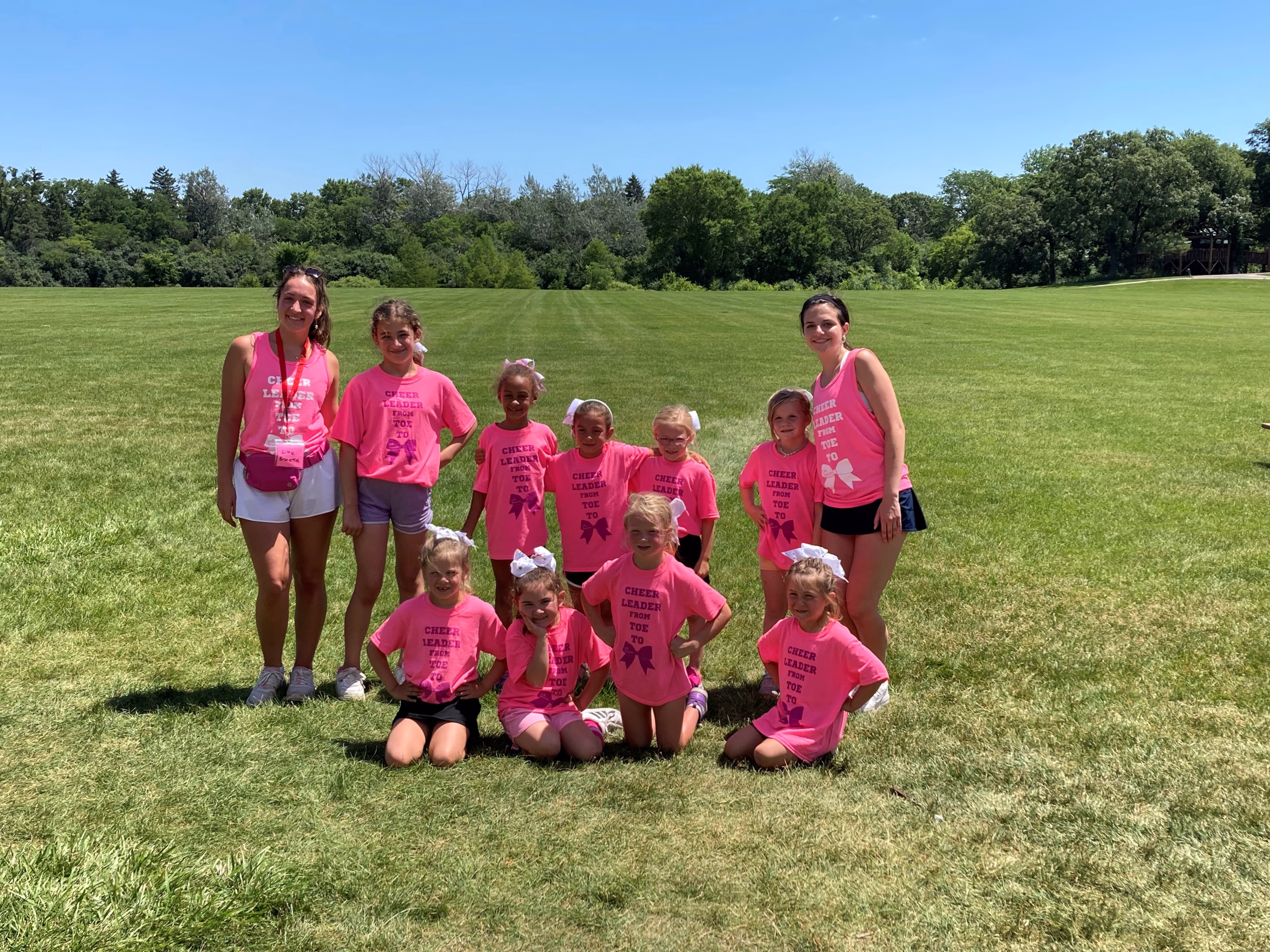 A group of young girls and two female coaches pose for the camera in two rows, in the middle of the field wearing their cheer t-shirts and white hair bows.