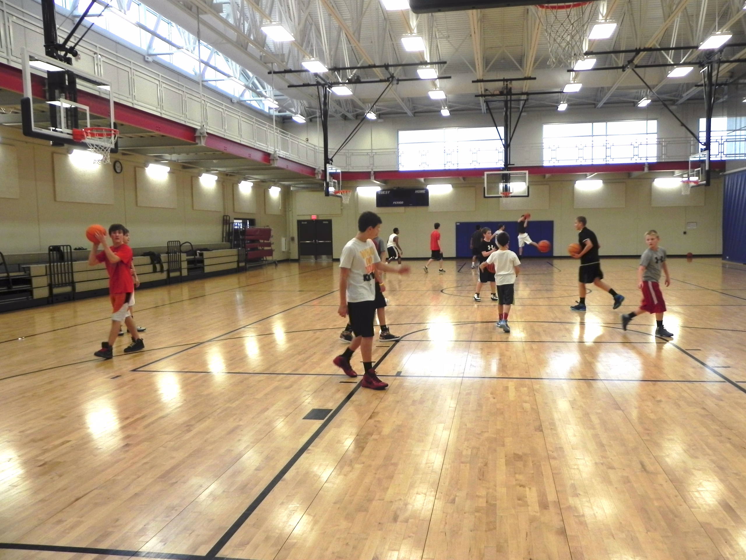 A group of children and a coach practice basketball inside a gym, dribbling and moving around the court under bright overhead lights.