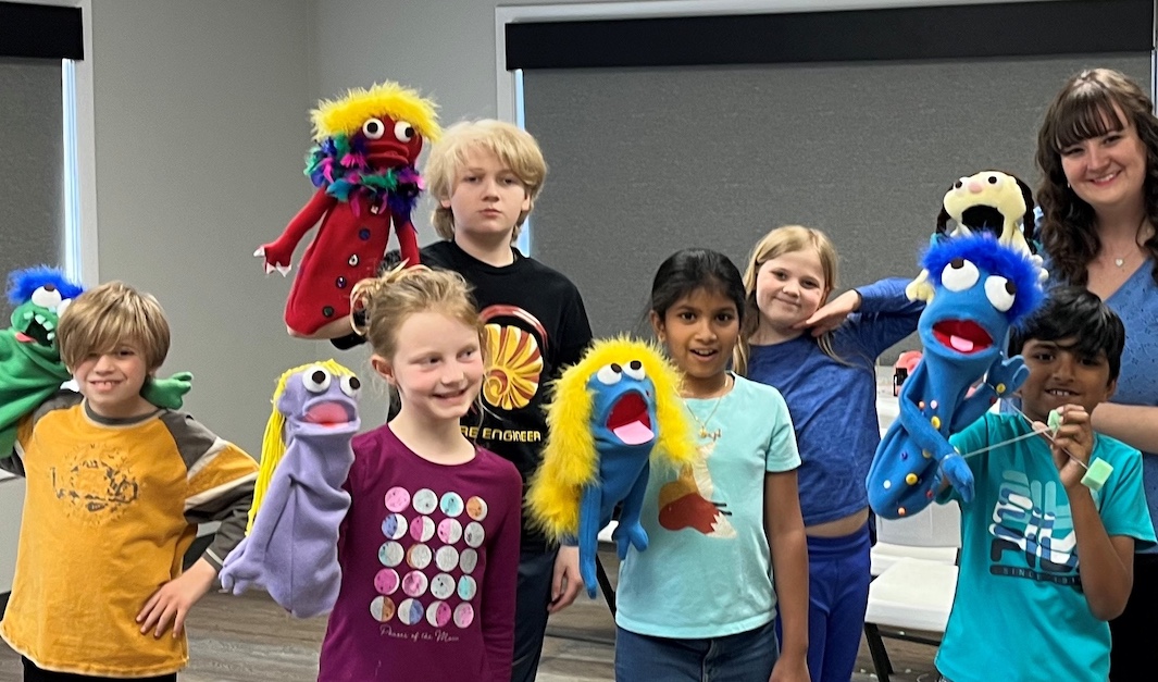 A group of children stand together holding colorful handmade puppets, smiling and posing in a classroom setting while an adult stands off to the side.