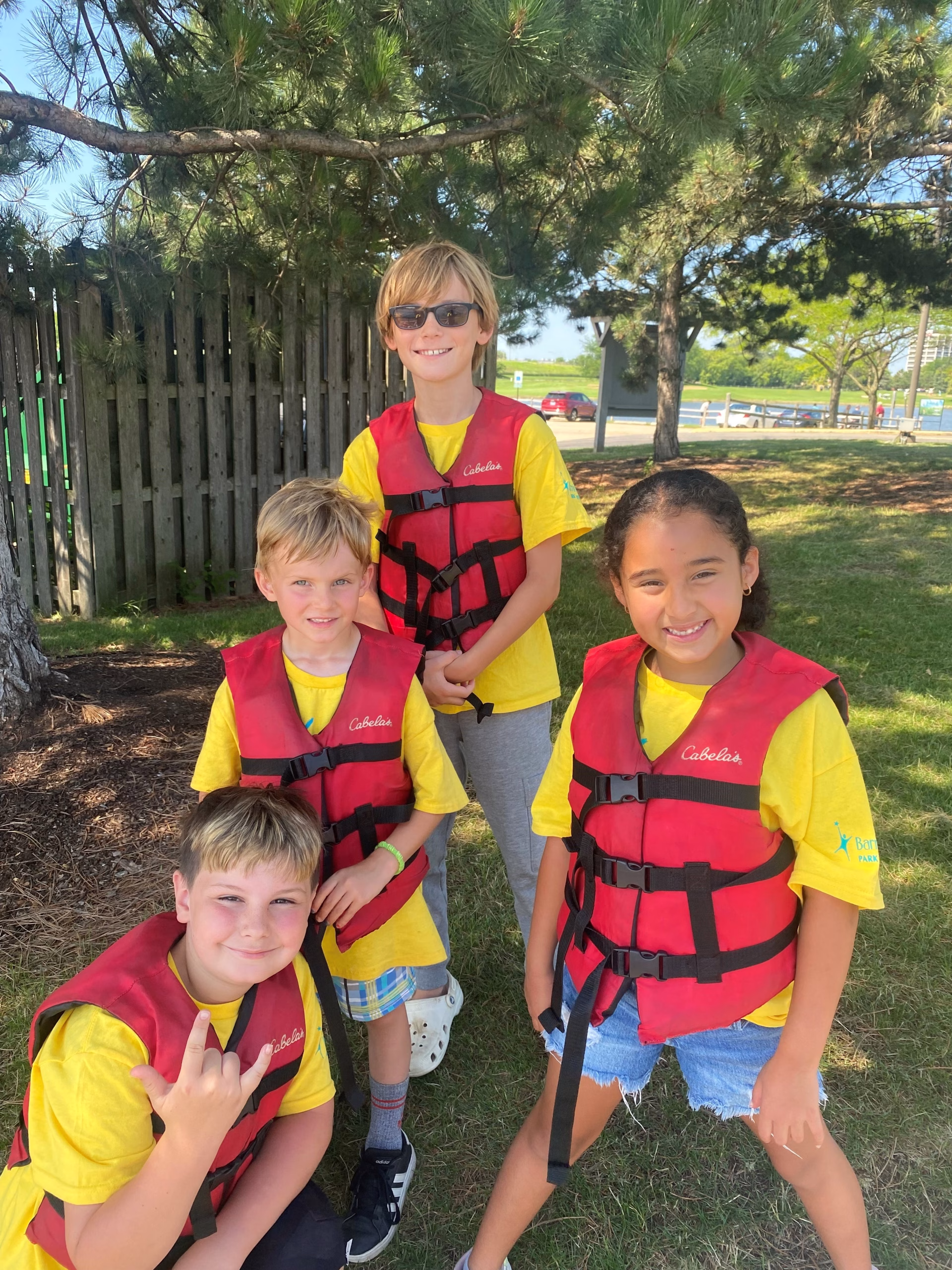 A group of four young children pose outdoors while wearing their yellow camp t-shirts and life jackets.